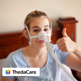Woman sitting in bed with CPAP mask on smiling and giving a thumbs up