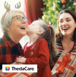 Little girl giving her grandma a kiss on the cheek and in the background little girl's mother smiling and holding a present 