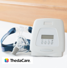 CPAP machine and mask on bed with white bedsheets and oak headboard