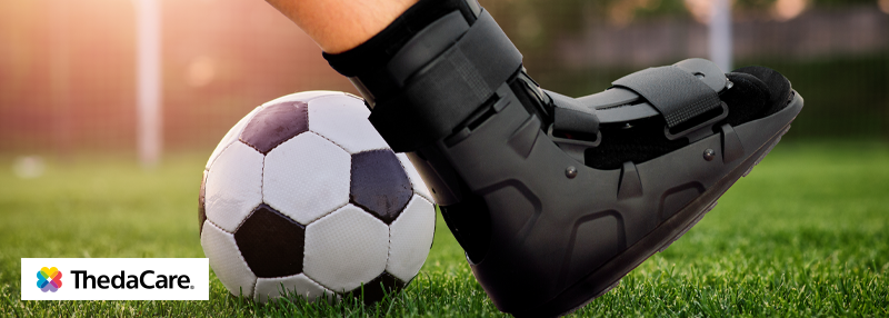 Foot in orthopedic boot next to soccer ball on soccer field.